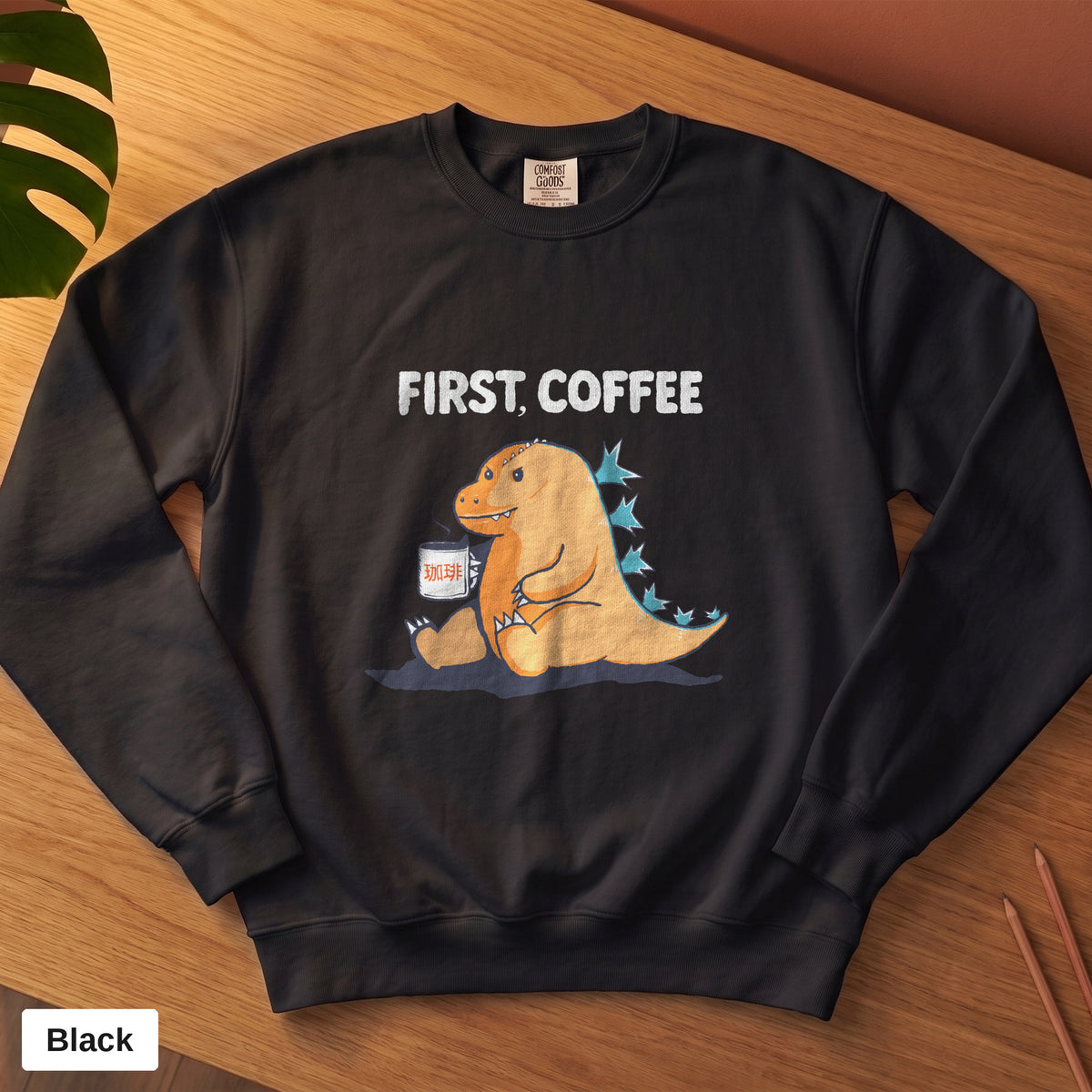 Black Comfort Colors crewneck sweatshirt, folded flat lay, First Coffee kaiju design printed on the chest