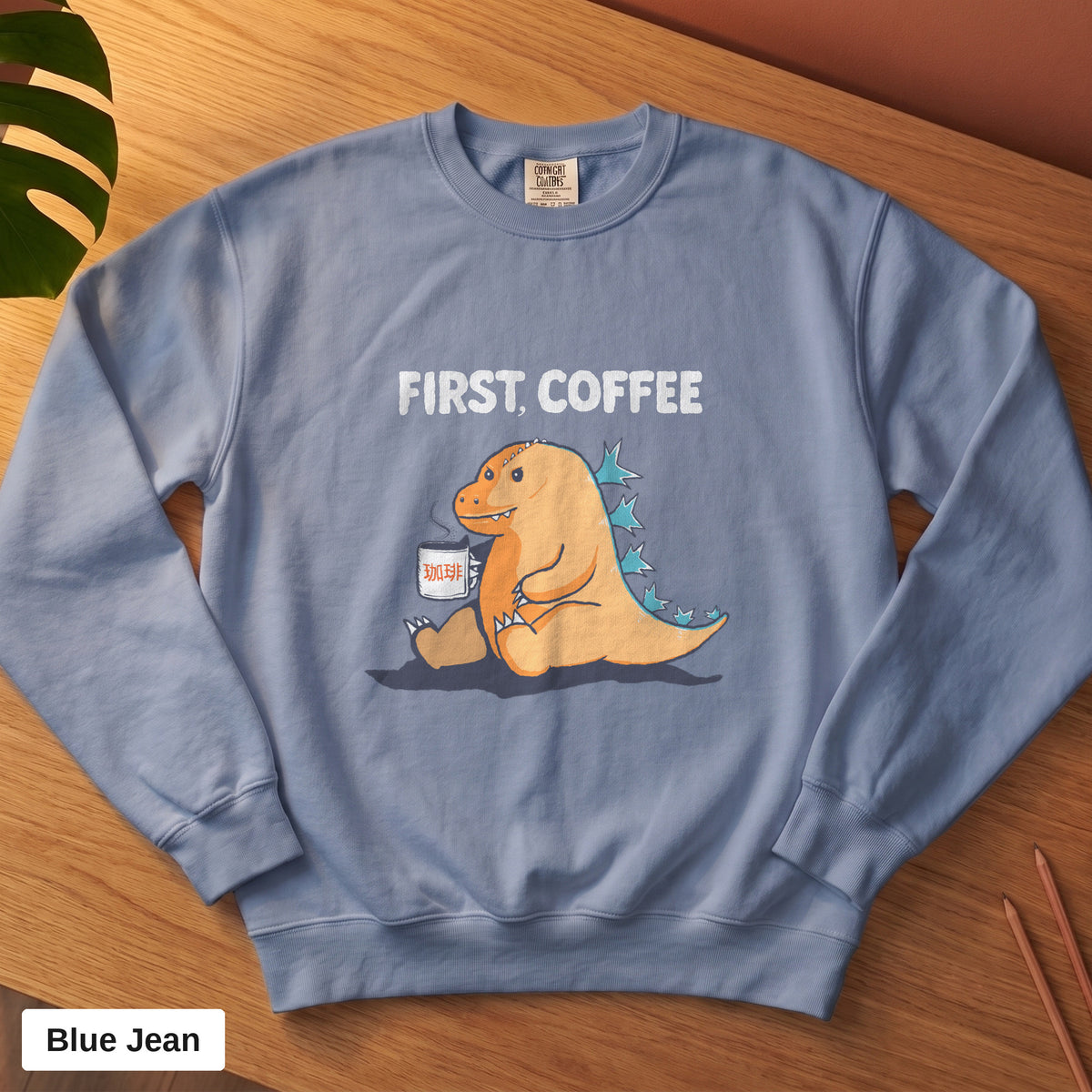 Blue Jean Comfort Colors crewneck sweatshirt, folded flat lay, First Coffee kaiju design printed on the chest
