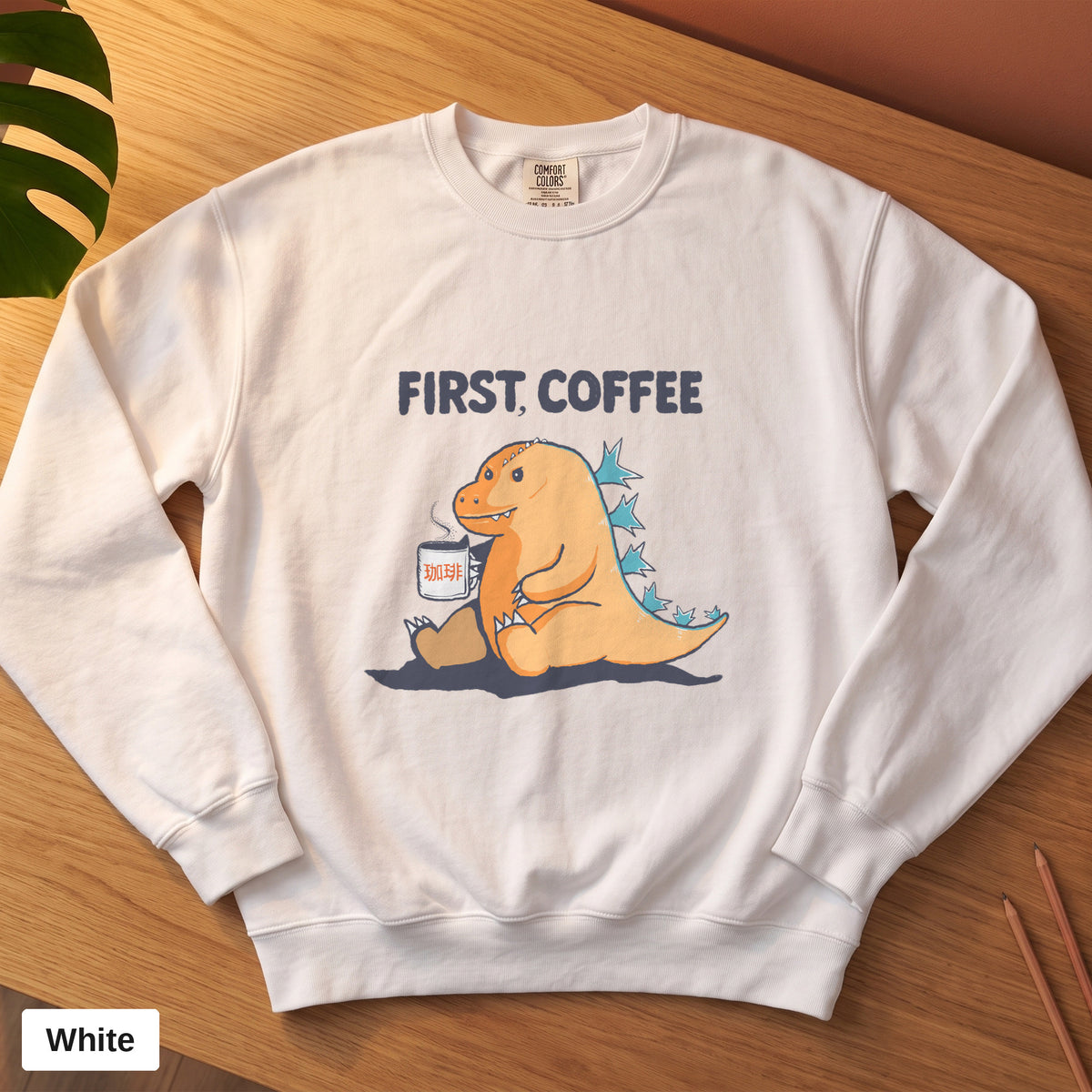 White Comfort Colors crewneck sweatshirt, folded flat lay, First Coffee kaiju design printed on the chest