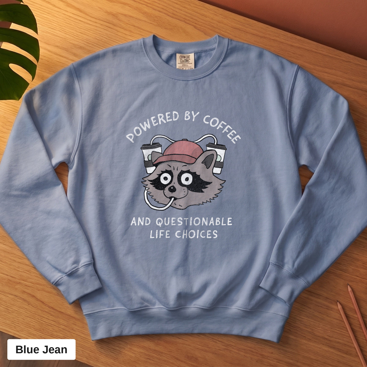 Blue Jean Comfort Colors crewneck sweatshirt, folded flat lay, Powered By Coffee And Questionable Life Choices raccoon design printed on the chest