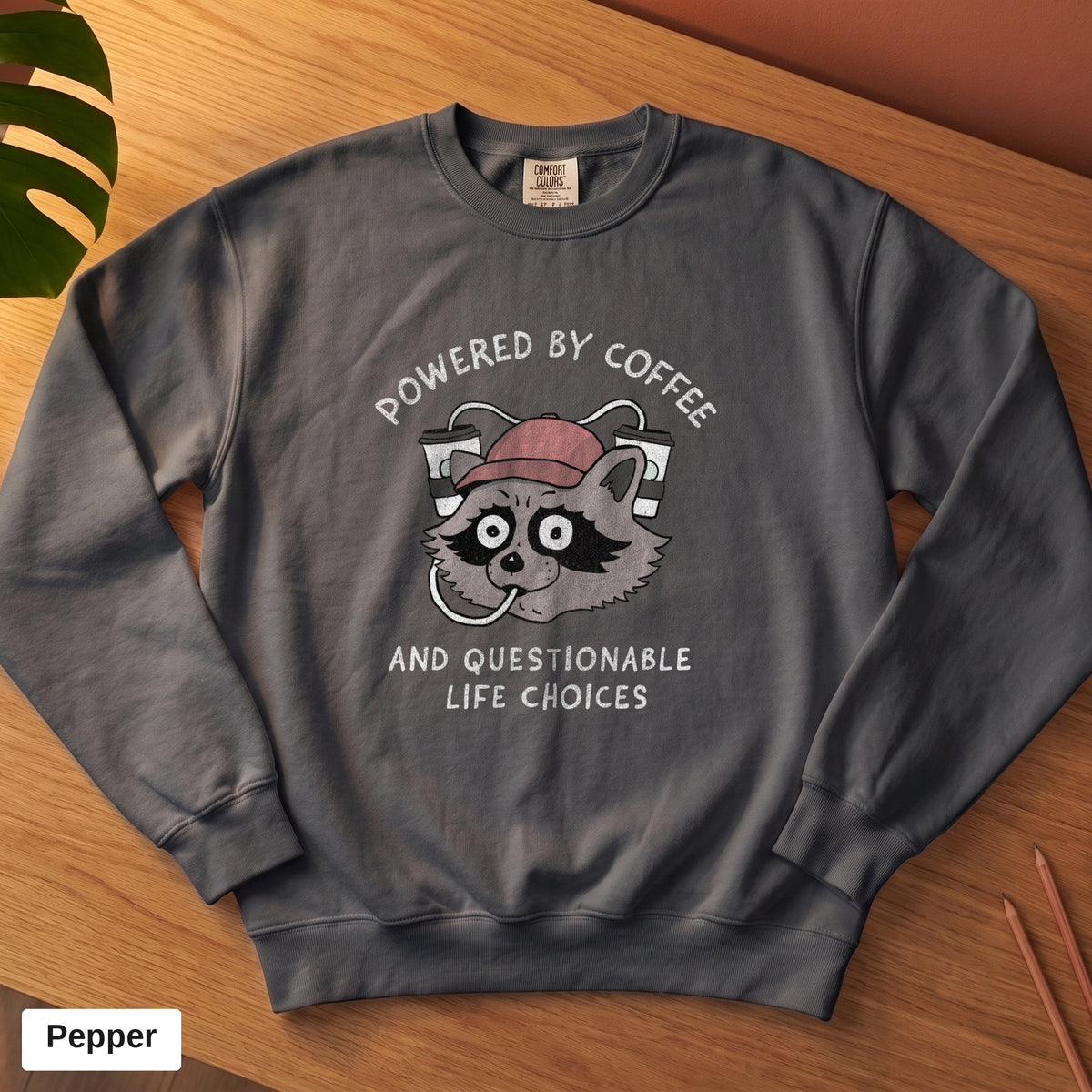 Pepper Comfort Colors crewneck sweatshirt, folded flat lay, Powered By Coffee And Questionable Life Choices raccoon design printed on the chest