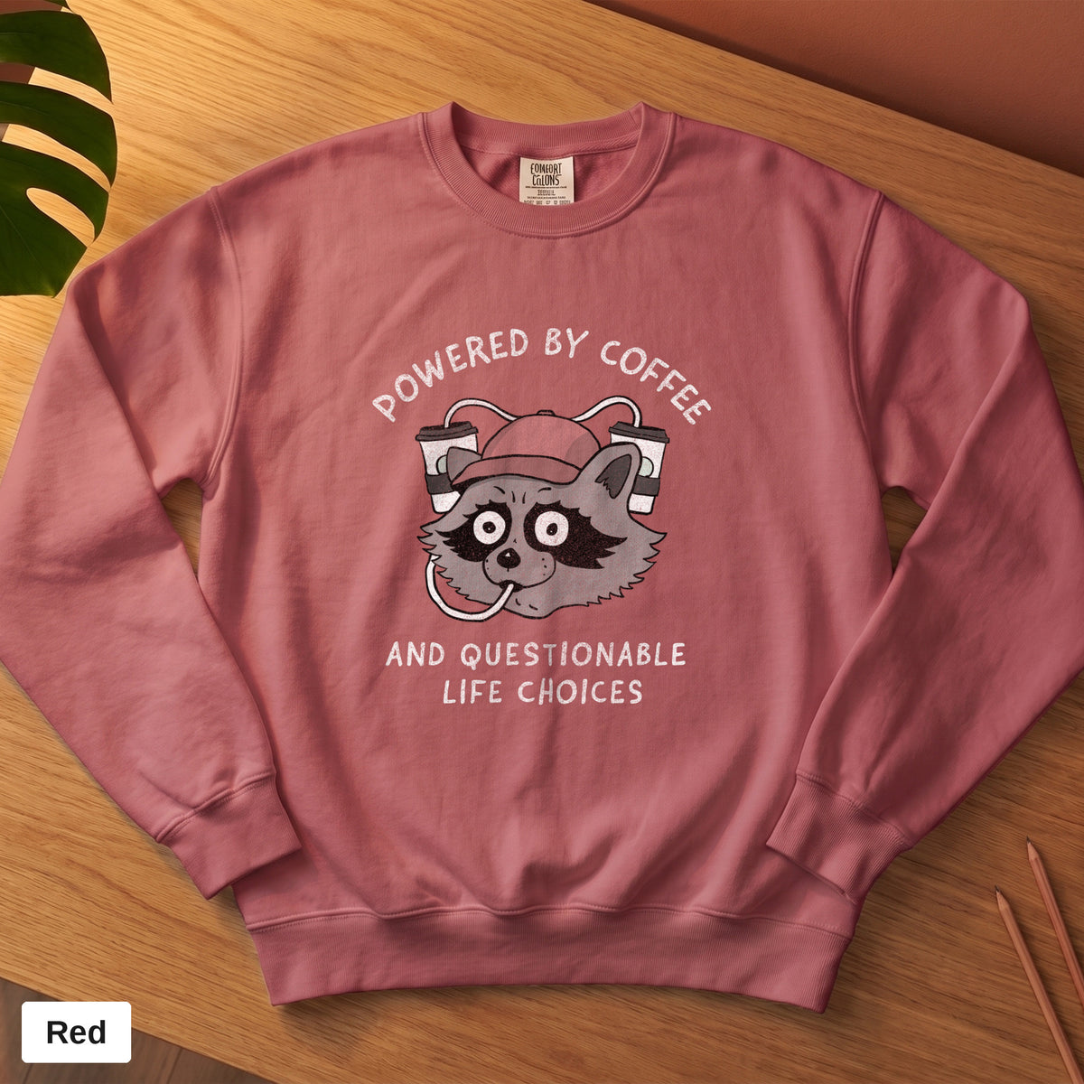 Crimson Comfort Colors crewneck sweatshirt, folded flat lay, Powered By Coffee And Questionable Life Choices raccoon design printed on the chest
