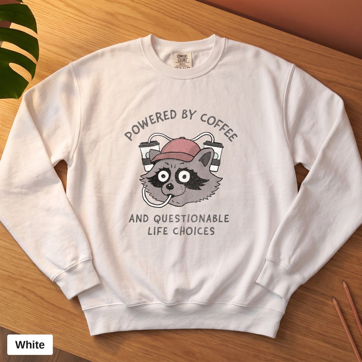 White Comfort Colors crewneck sweatshirt, folded flat lay, Powered By Coffee And Questionable Life Choices raccoon design printed on the chest