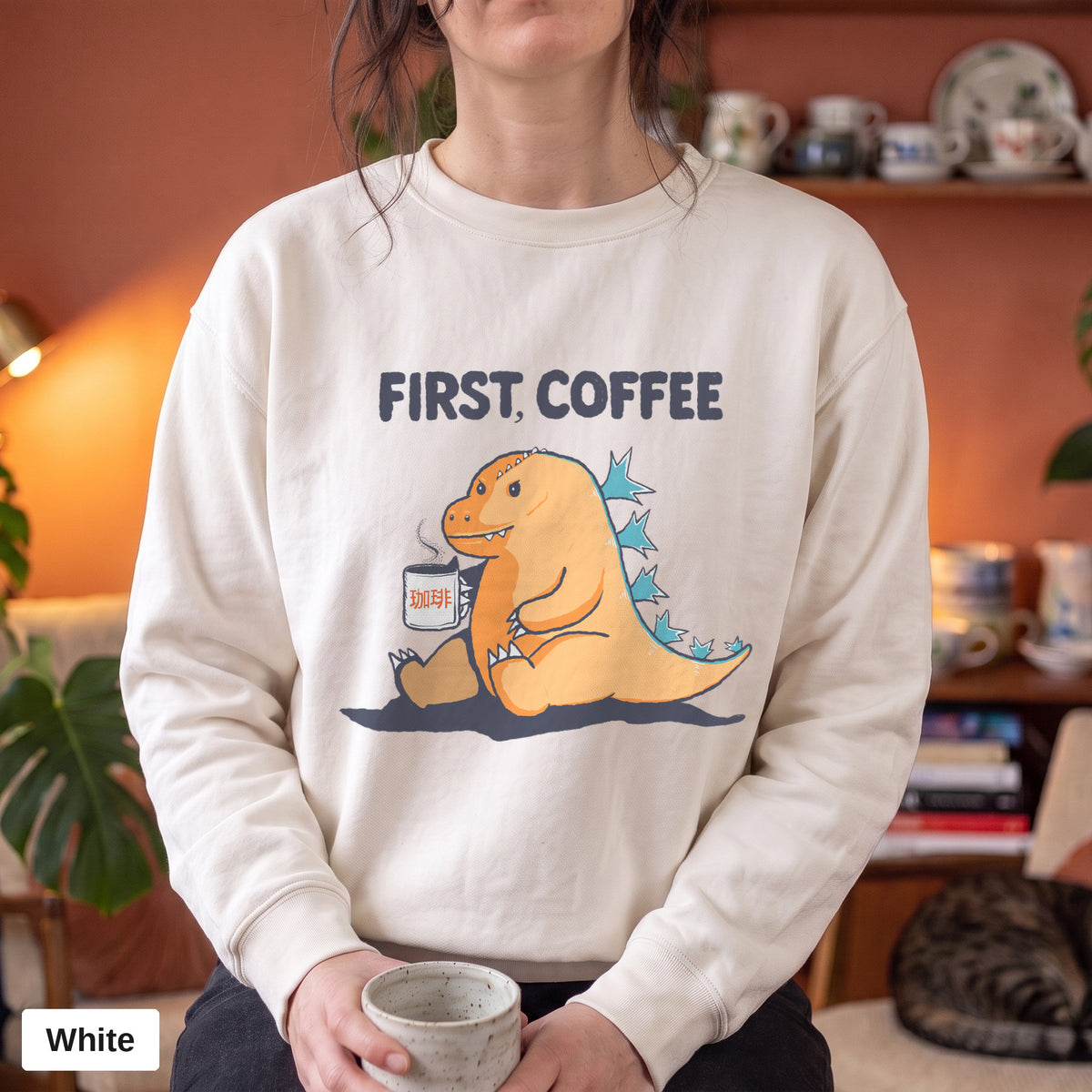 White Comfort Colors crewneck sweatshirt worn by a model, First Coffee kaiju design printed on the chest