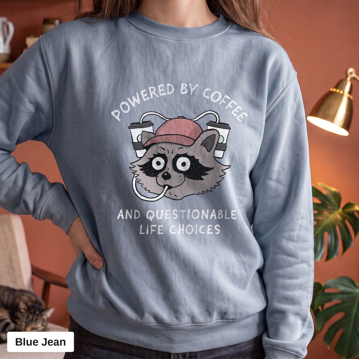 Blue Jean Comfort Colors crewneck sweatshirt worn by a model, Powered By Coffee And Questionable Life Choices raccoon design printed on the chest